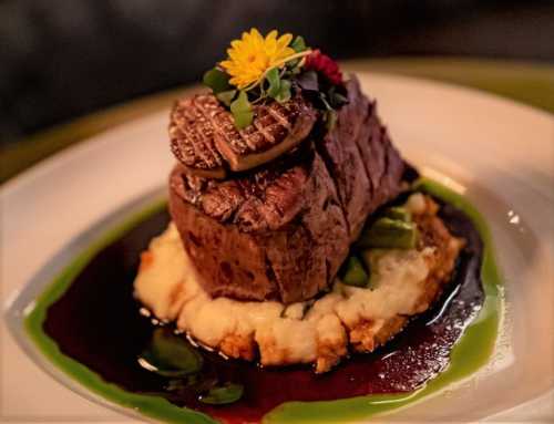 A gourmet dish featuring a filet mignon topped with foie gras, served on creamy mashed potatoes with a rich sauce and garnishes.