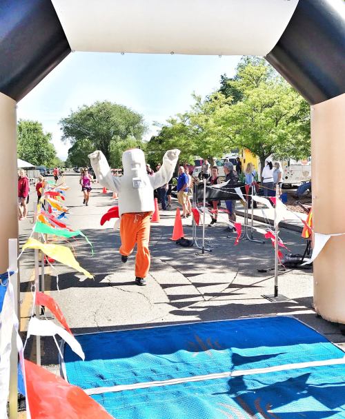 A person in a large mascot costume runs through a finish line arch at a lively outdoor event.