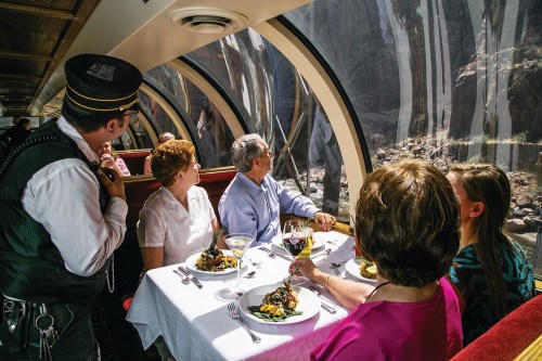 A group dining on a train, enjoying meals and drinks while admiring scenic views through large windows.