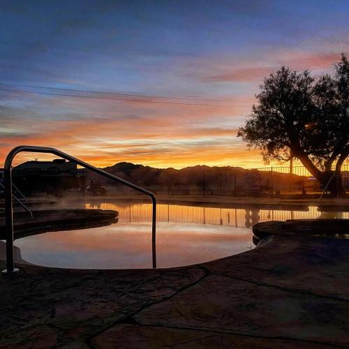 A serene hot spring at sunset, with steam rising and mountains silhouetted against a colorful sky.