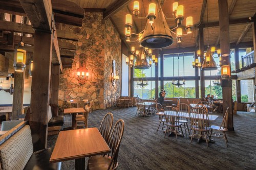 Cozy restaurant interior with wooden beams, stone walls, and large windows overlooking greenery. Warm lighting and seating.