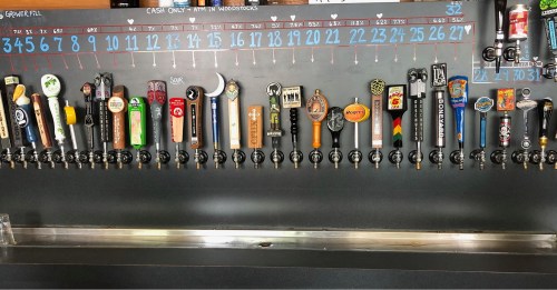 A wall-mounted beer tap system with various colorful tap handles and a menu board above listing beer options.