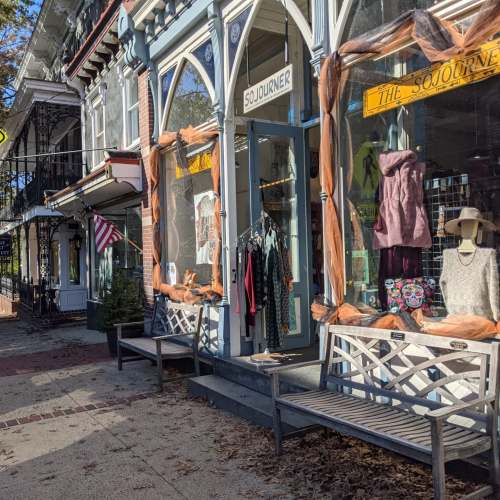 A charming storefront with large windows displaying clothing, flanked by benches and American flags on a sunny day.
