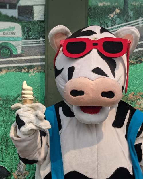 A costumed cow character wearing red sunglasses holds an ice cream cone, standing in front of a colorful mural.