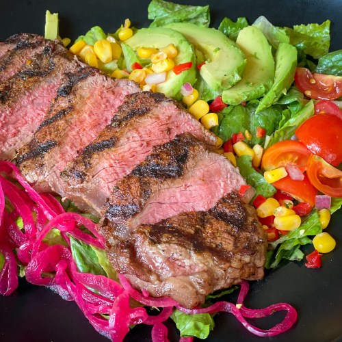 Grilled steak on a bed of greens, topped with avocado, corn, cherry tomatoes, and pickled onions.