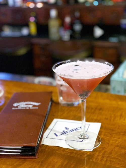 A pink cocktail in a martini glass sits on a wooden bar, next to a menu and a napkin.