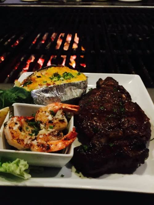 A plate featuring grilled shrimp, a baked potato, and a juicy steak, with a grill in the background.