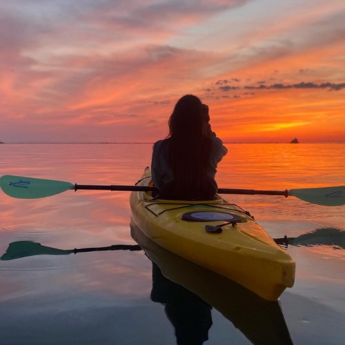 A person in a yellow kayak gazes at a vibrant sunset over calm waters, reflecting the colorful sky.
