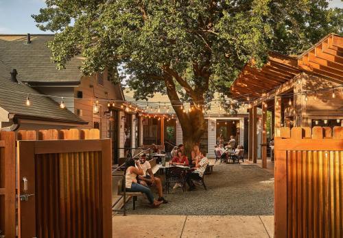A cozy outdoor space with people dining under a large tree, surrounded by wooden structures and string lights.