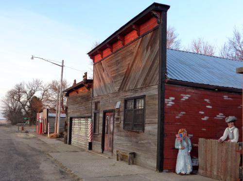 A rustic street scene featuring weathered wooden buildings and a vintage Americana figure holding a flag.