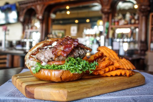 A juicy burger with bacon and lettuce on a bun, served with sweet potato fries on a wooden board.