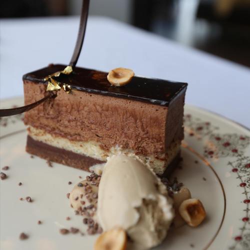 A decadent chocolate mousse cake with a glossy glaze, garnished with hazelnuts and a scoop of creamy ice cream.