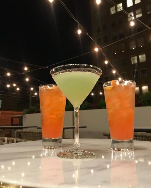 Three colorful cocktails on a marble table, illuminated by string lights at night.
