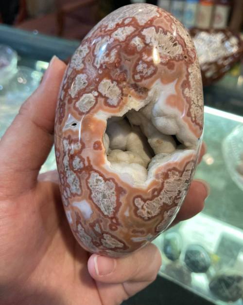 A hand holding a polished, oval-shaped stone with intricate patterns and a hollow center revealing gray crystals inside.