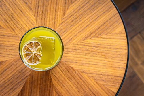 A vibrant yellow drink with a dried lime slice sits on a wooden table with a starburst pattern.