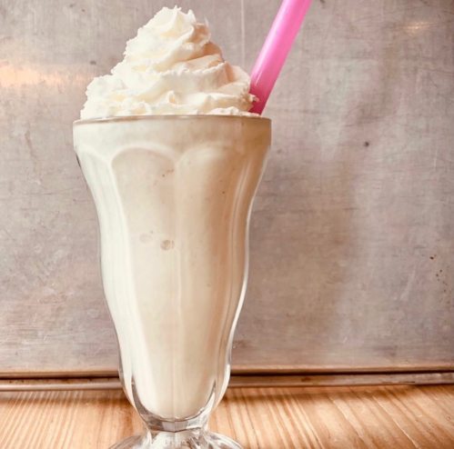 A tall glass of creamy milkshake topped with whipped cream and a pink straw, set against a textured background.