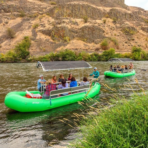 Two green rafts on a river with people enjoying a scenic float, surrounded by rocky hills and greenery.