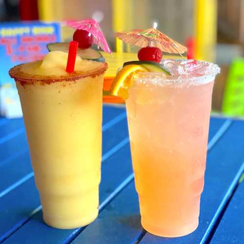 Two colorful cocktails with umbrellas, garnished with fruit, on a blue table. One is yellow and the other is pink.