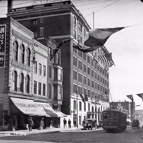 Historic street scene from the 1930s featuring buildings, a streetcar, and flags hanging overhead.