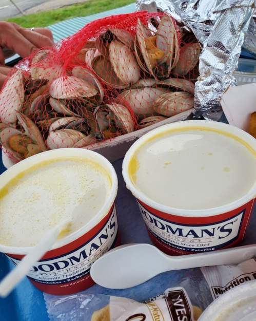 Two cups of clam chowder sit beside a netted bag of clams, with a foil-wrapped item in the background.
