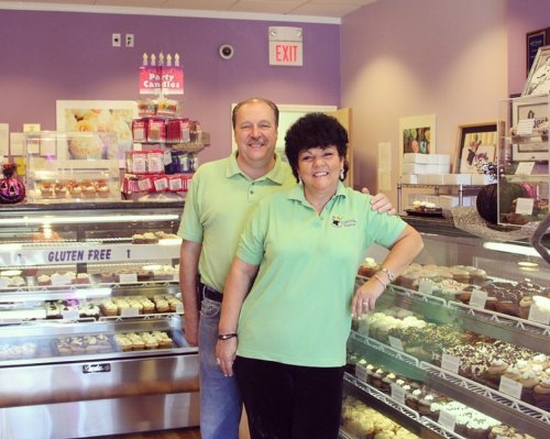 A man and woman stand smiling in a bakery, surrounded by displays of cupcakes and pastries.