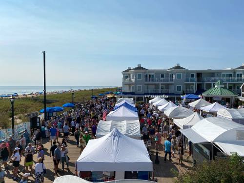 A bustling beachside market with white tents, crowds of people, and a view of the ocean in the background.