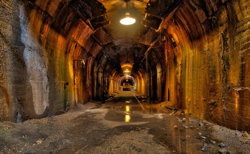 A dimly lit underground tunnel with wet floors and textured walls, creating an eerie, atmospheric scene.