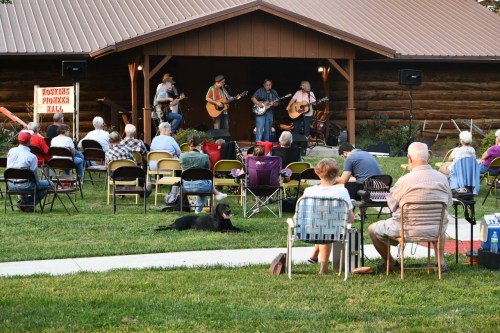 A live band performs on stage for an audience seated outdoors, with a dog resting nearby.