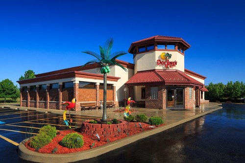 Exterior of a restaurant with a palm tree, colorful decor, and a clear blue sky. Parking lot in front.