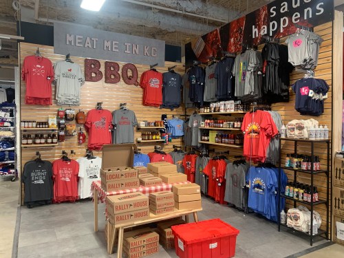 A display of BBQ-themed merchandise, including shirts and sauces, in a retail space with wooden walls and stacked boxes.
