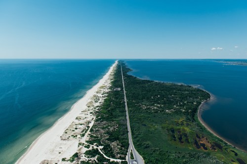Aerial view of a long, narrow strip of land bordered by ocean on one side and a bay on the other, under a clear blue sky.