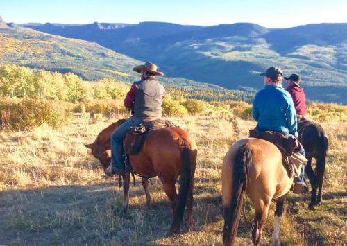 Three horseback riders view a scenic mountain landscape during sunset, surrounded by autumn foliage.