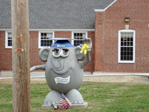 A gray elephant statue wearing a graduation cap, holding a yellow ribbon, in front of a brick building.