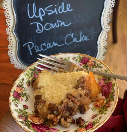 A slice of upside-down pecan cake on a floral plate, with a fork beside it and a chalkboard sign in the background.