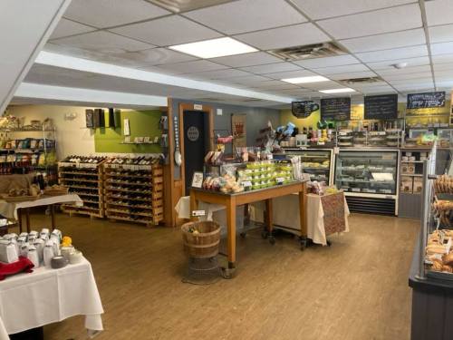 A cozy market interior featuring shelves of wine, baked goods, and a display case, with tables set for customers.