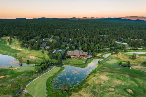 Aerial view of a golf course surrounded by trees and water, with mountains in the background at sunset.
