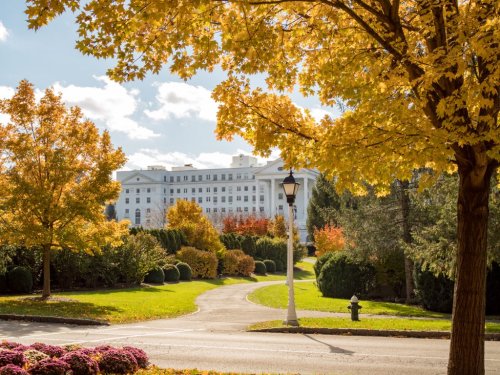 A scenic view of a park with vibrant autumn trees and a white building in the background under a clear blue sky.