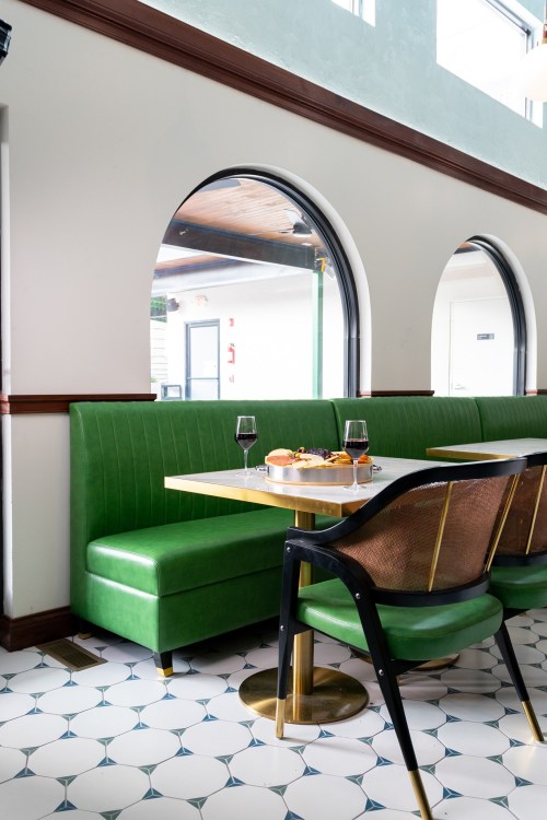 A cozy restaurant corner with a green booth, a table set with drinks and pastries, and large arched windows.