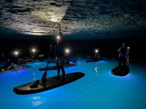 A group of people on paddleboards with headlamps, exploring a dark cave with glowing blue water.