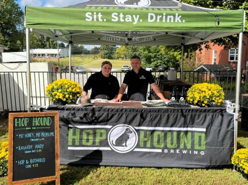 A brewing booth with two staff members, yellow flowers, and a chalkboard menu at an outdoor event.