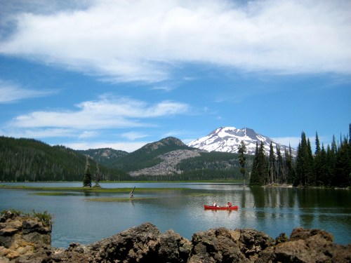 A serene lake surrounded by mountains and trees, with two people kayaking under a blue sky with wispy clouds.