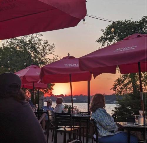 A sunset view over a lake, framed by red umbrellas at an outdoor restaurant with diners enjoying the evening.