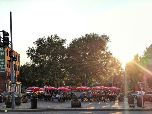 Outdoor dining scene at sunset, with people seated under red umbrellas in a lively urban setting.