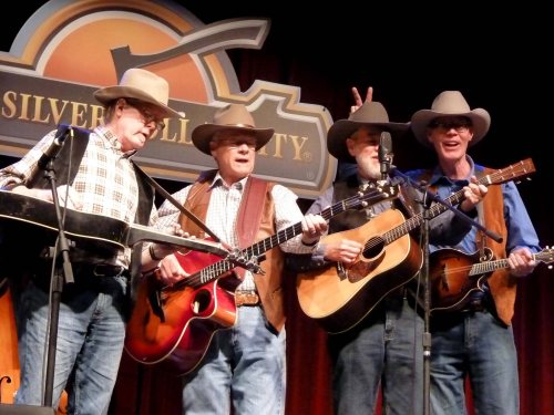 Four musicians in cowboy hats perform on stage, playing guitars and a steel guitar, with a playful gesture from one.