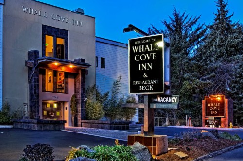 Exterior of Whale Cove Inn at dusk, featuring a welcoming sign and a "Vacancy" notice. Lush greenery surrounds the entrance.