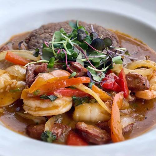 A colorful dish featuring shrimp, sausage, and vegetables in a savory sauce, garnished with microgreens.