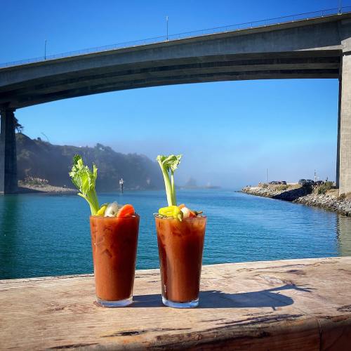 Two Bloody Mary cocktails with celery and vegetables, set against a scenic waterfront and bridge under a clear blue sky.