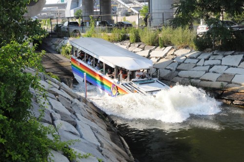A colorful boat splashes through a waterway, surrounded by greenery and urban structures.