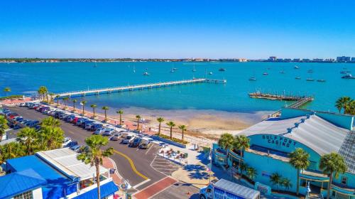 Aerial view of a sunny waterfront with a pier, boats, and palm trees, alongside a vibrant coastal road.