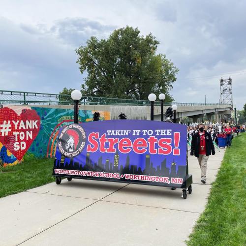 A parade with a banner reading "Takin' it to the Streets!" in Worthington, MN, with people walking on a path.